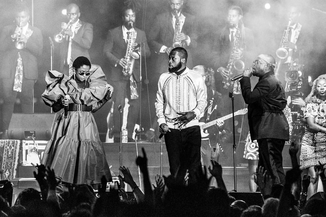 Live concert scene featuring three singers on stage with a band, bathed in monochrome lighting, performing before a crowd.