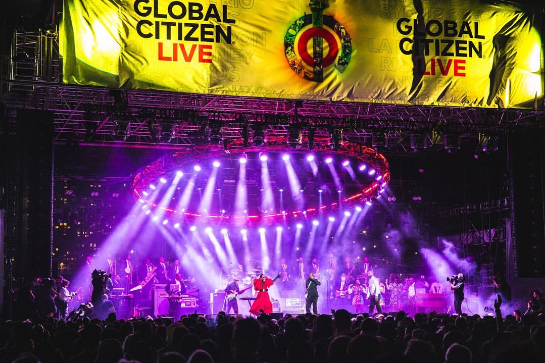 A concert stage featuring performers under bright lights, with a large crowd visible in the foreground and a 'Global Citizen Live' banner overhead.
