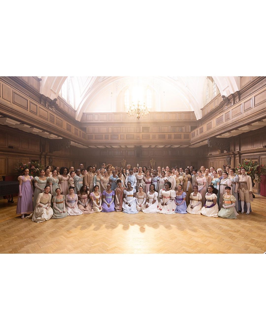 Large group of people gathered indoors on a wooden floor with elegant architecture. People are in dresses and formal wear.