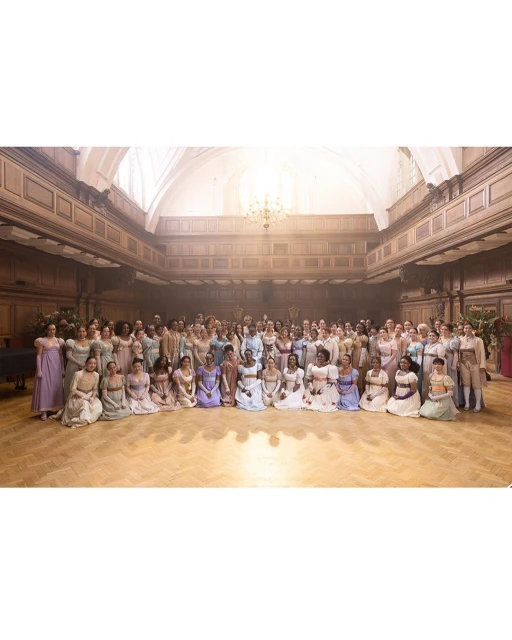 Large group of people gathered indoors on a wooden floor with elegant architecture. People are in dresses and formal wear.