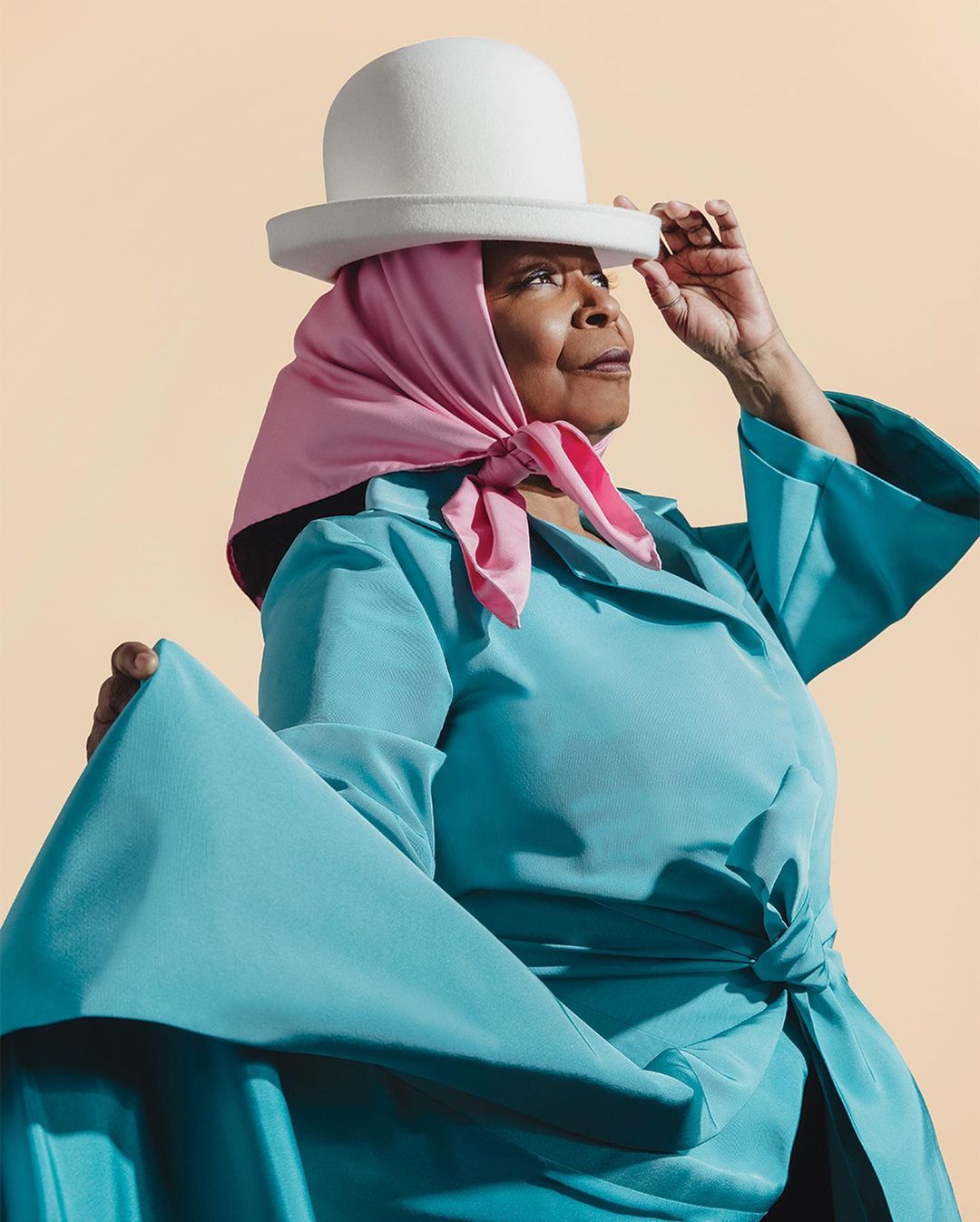 Portrait of a mature Black woman in a teal dress and white hat, headscarf, posing in a studio setting with a solid light-peach backdrop.
