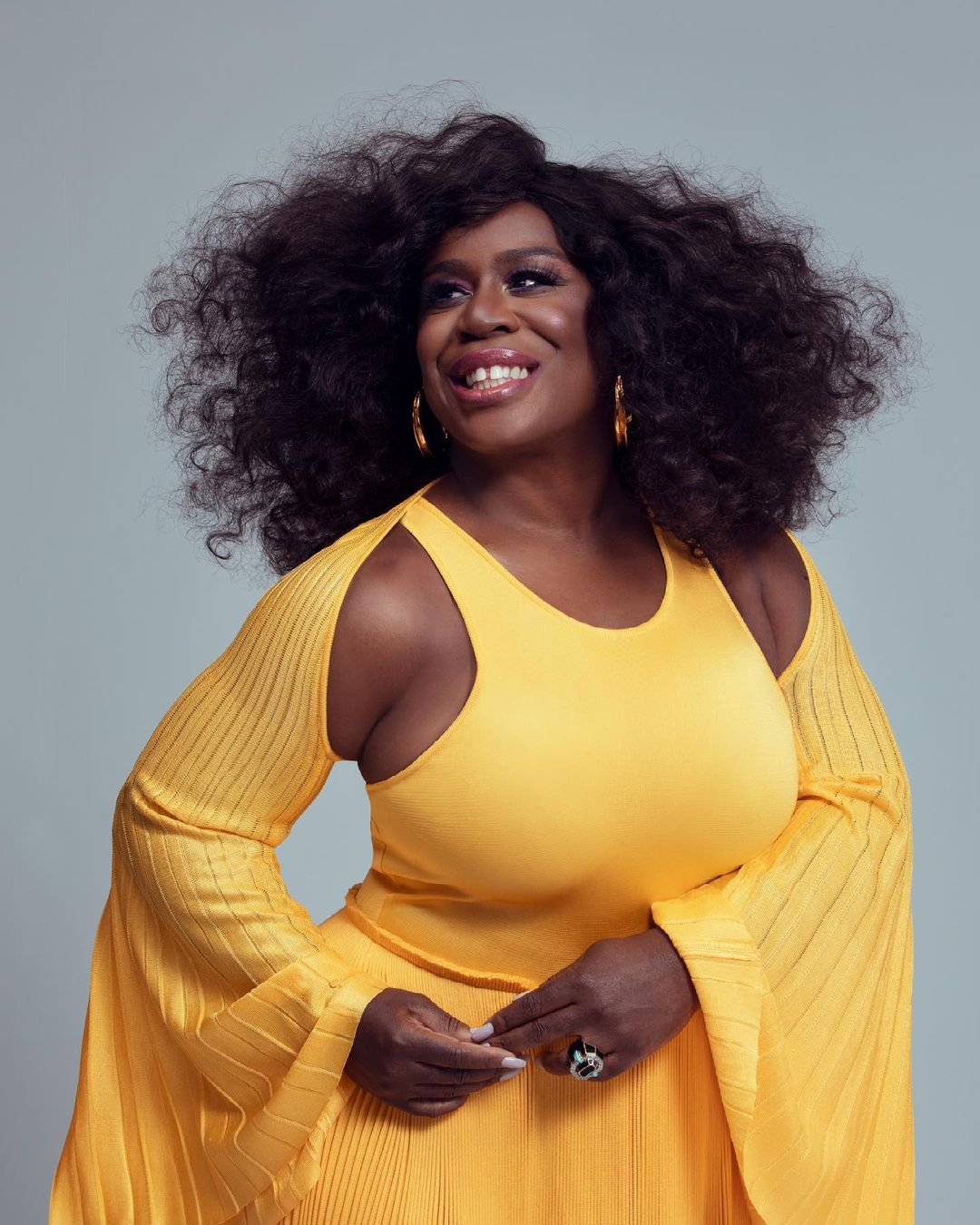 Portrait of a smiling African-American woman with curly dark hair, wearing a yellow dress, hoop earrings, and a ring, posing indoors.