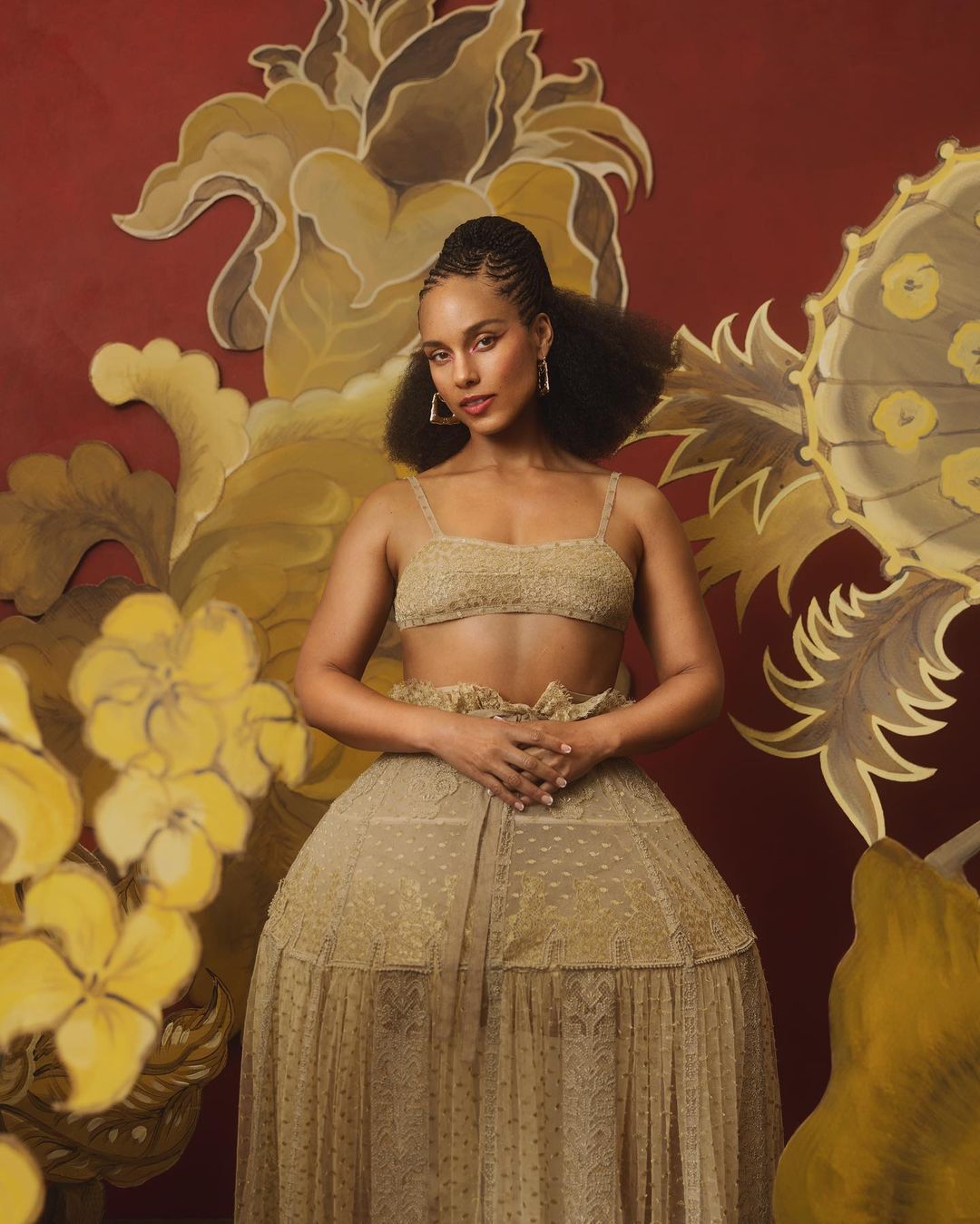Portrait of Alicia Keys wearing a gold two-piece outfit against a backdrop of stylized flowers in shades of gold and red.