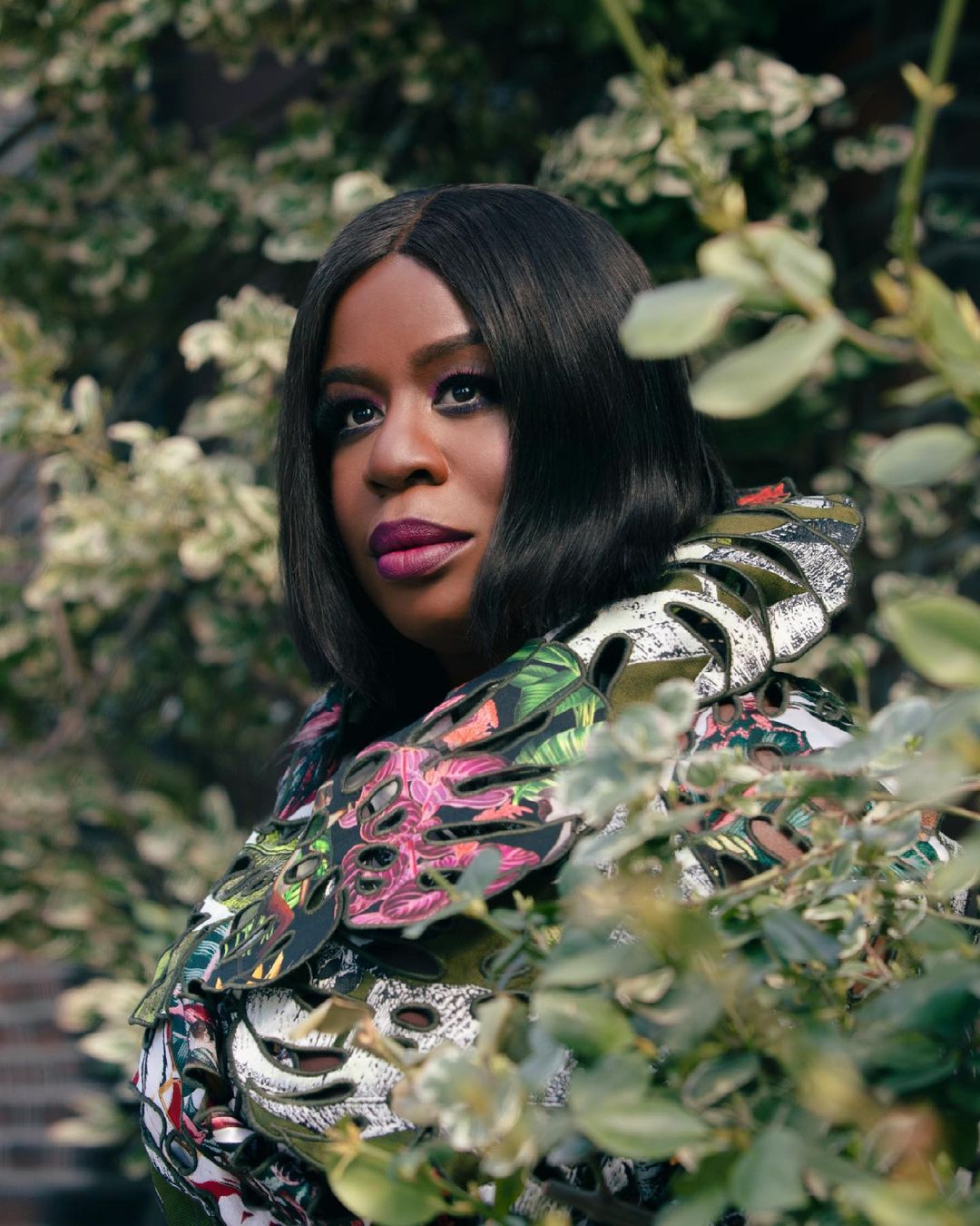 Portrait of Black woman with short black hair and bold lipstick, wearing a floral dress, posing outdoors surrounded by leafy plants.