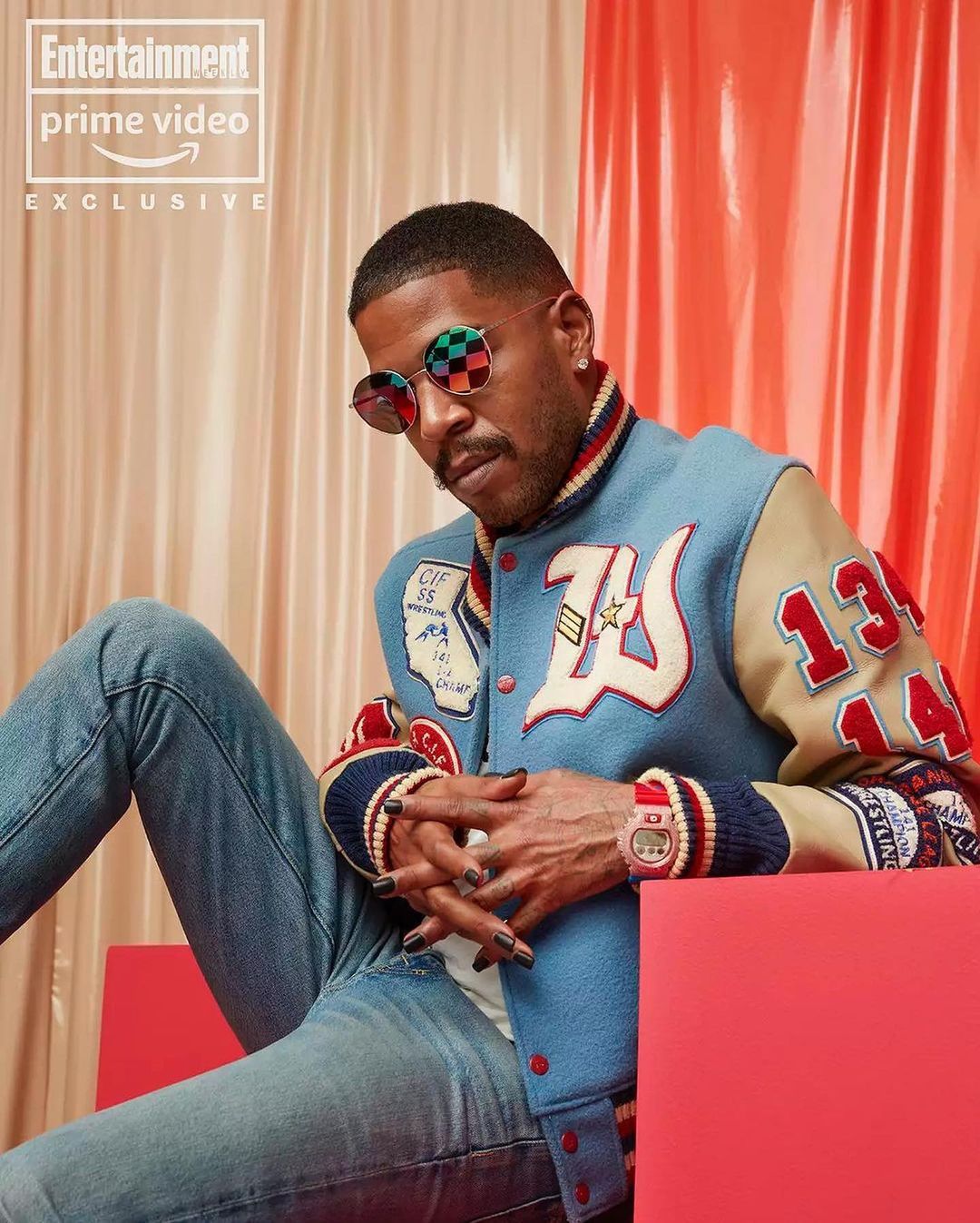 Adult male seated, wearing letterman jacket and sunglasses with a peach and orange studio background for entertainment promo.