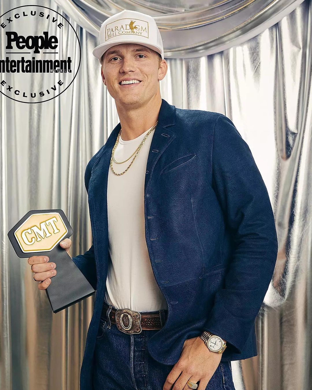 Young man in a blazer and ball cap holds a CMT award against a silver backdrop, wearing jeans and a decorative belt buckle.