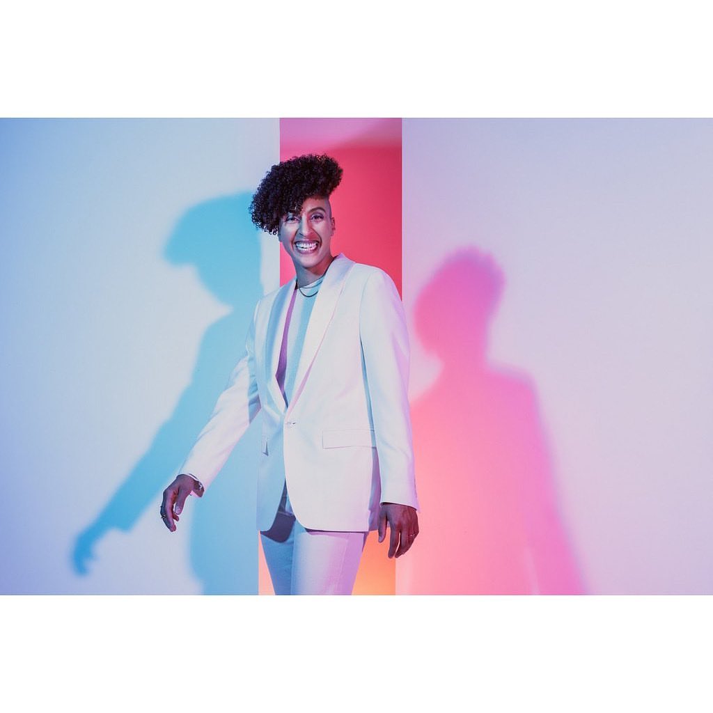 A person in a white suit smiles in front of a vibrant backdrop with a blend of pink, white, and blue hues, captured in a studio setting.