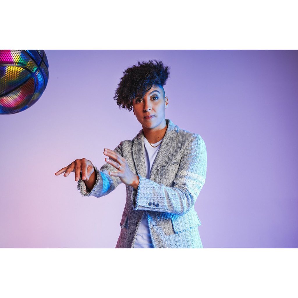 A woman in a gray patterned blazer extends her hands with a basketball hovering nearby against a purple backdrop in a studio shot.