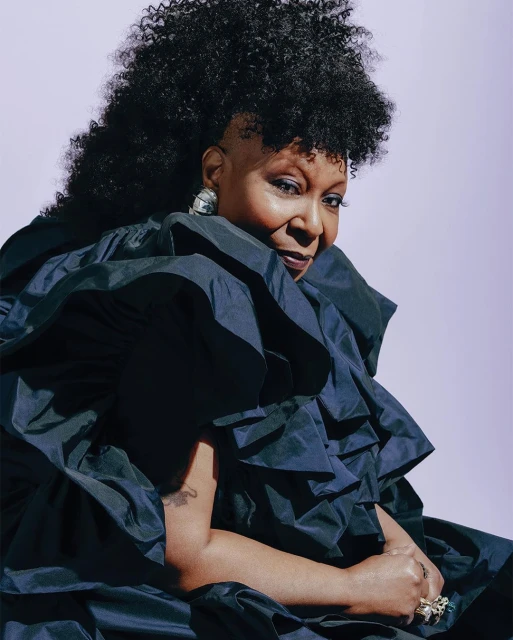 Portrait of a woman with voluminous dark curly hair wearing a voluminous black ruffled collar dress against a pale background