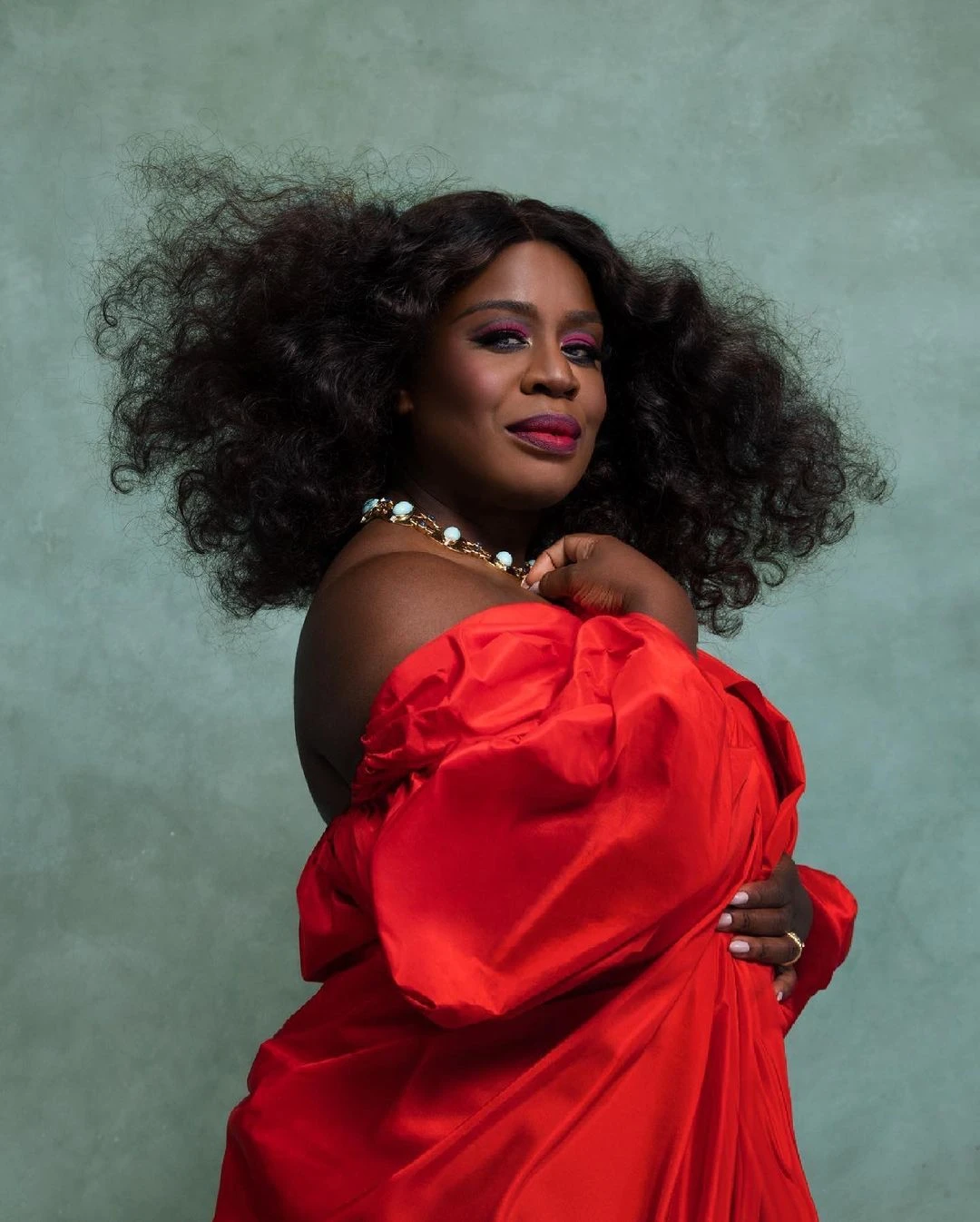 Portrait of a Black woman with voluminous curly hair wearing an off-the-shoulder red dress against a muted green backdrop.