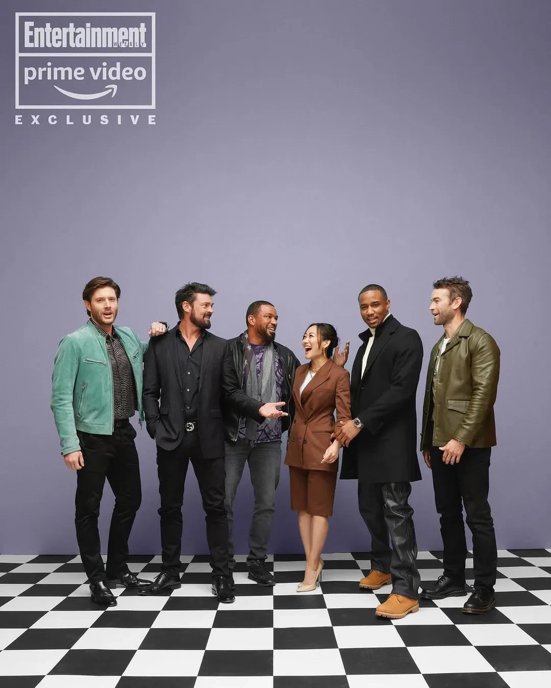 Group portrait of six actors standing together on a black and white checkerboard floor against a pale purple backdrop.