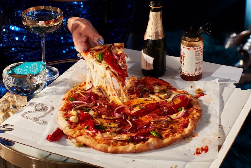 Close-up of pizza with toppings on a table with a glass of wine, champagne, a hot sauce bottle, and a key holder.