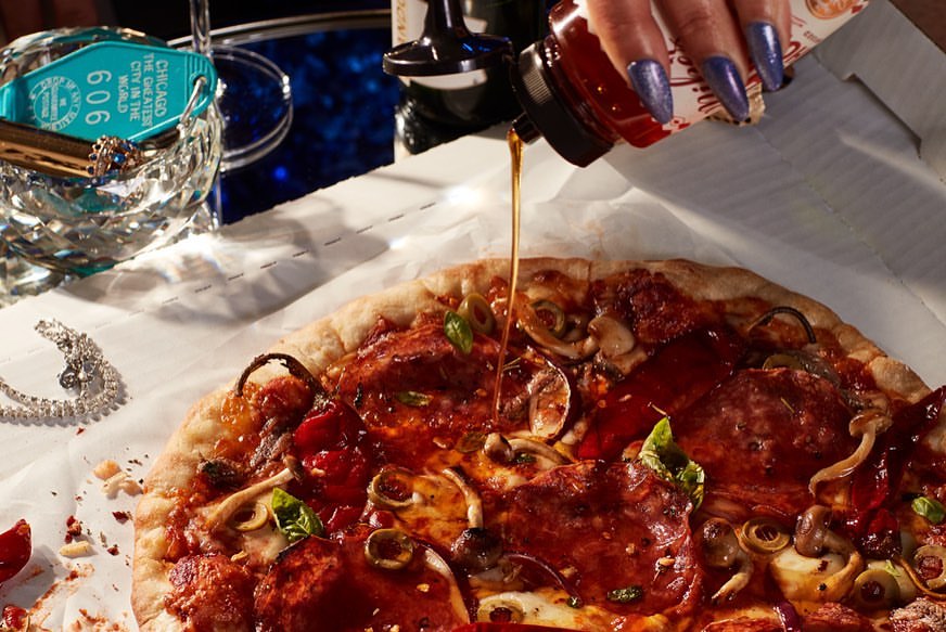 Overhead shot of artisan pizza drizzled with chili oil, showcasing culinary luxury with vibrant toppings on rustic crust and polished accessories.