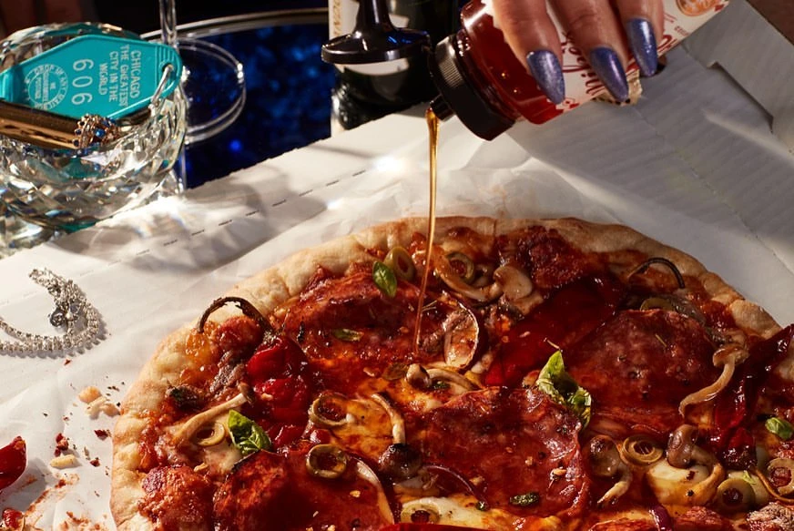 Overhead shot of artisan pizza drizzled with chili oil, showcasing culinary luxury with vibrant toppings on rustic crust and polished accessories.