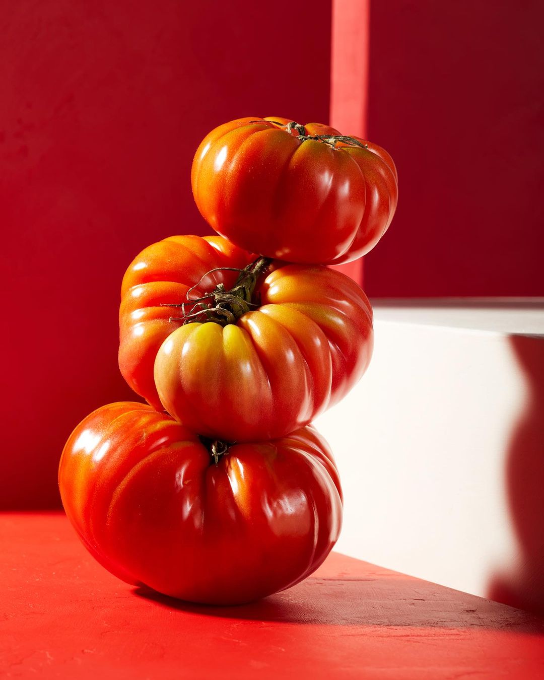 Still life shot of a stack of three ripe heirloom tomatoes against a vibrant red background with minimalist white architectural accents.