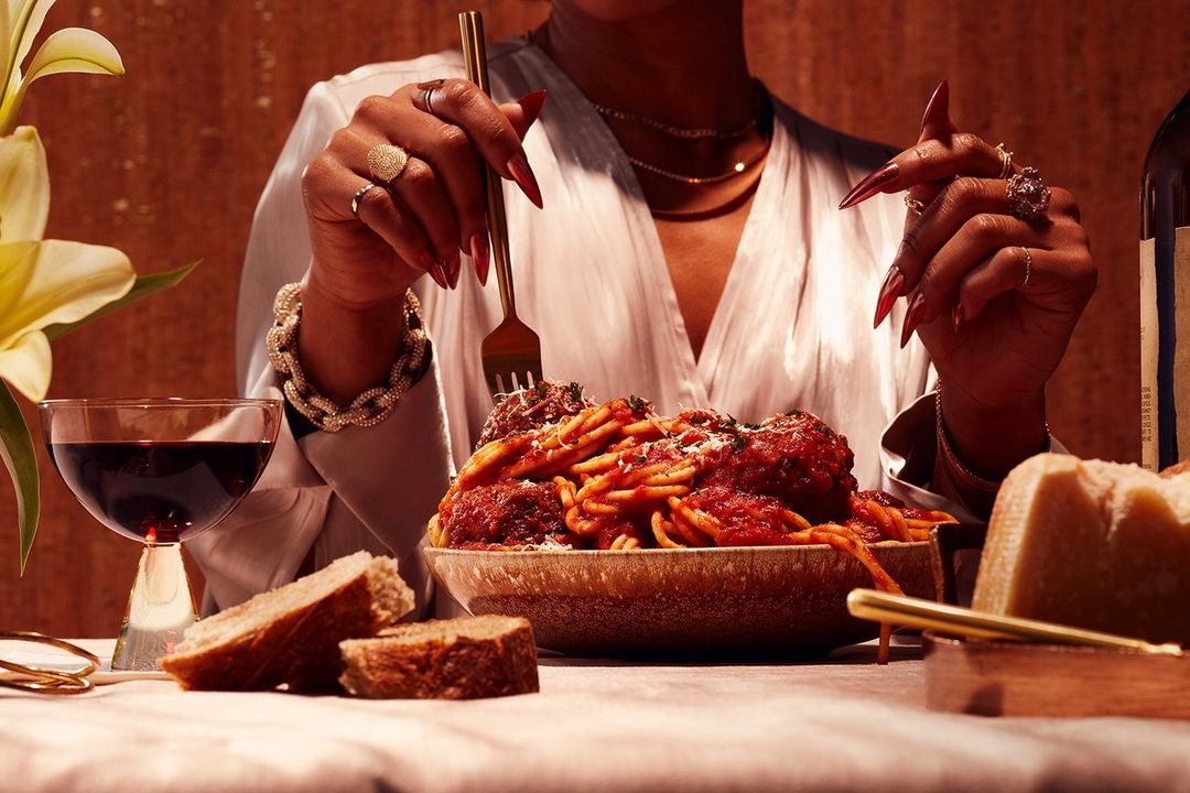 Woman with long red nails dines on spaghetti and meatballs with red wine and bread, set in warm, inviting, and elegant environment.