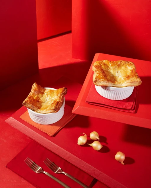 Overhead shot of two golden-crusted pot pies in ramekins on a red background, accompanied by forks and pearl onions.