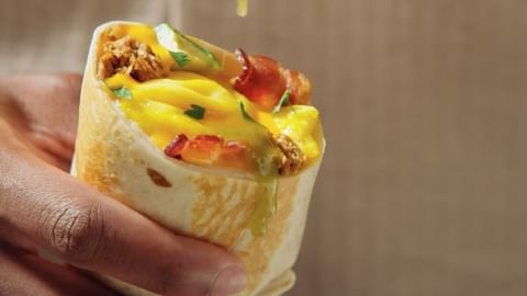 Close-up of a hand holding a breakfast burrito filled with eggs, meat, and vegetables, drizzled with sauce, against a blurred background.