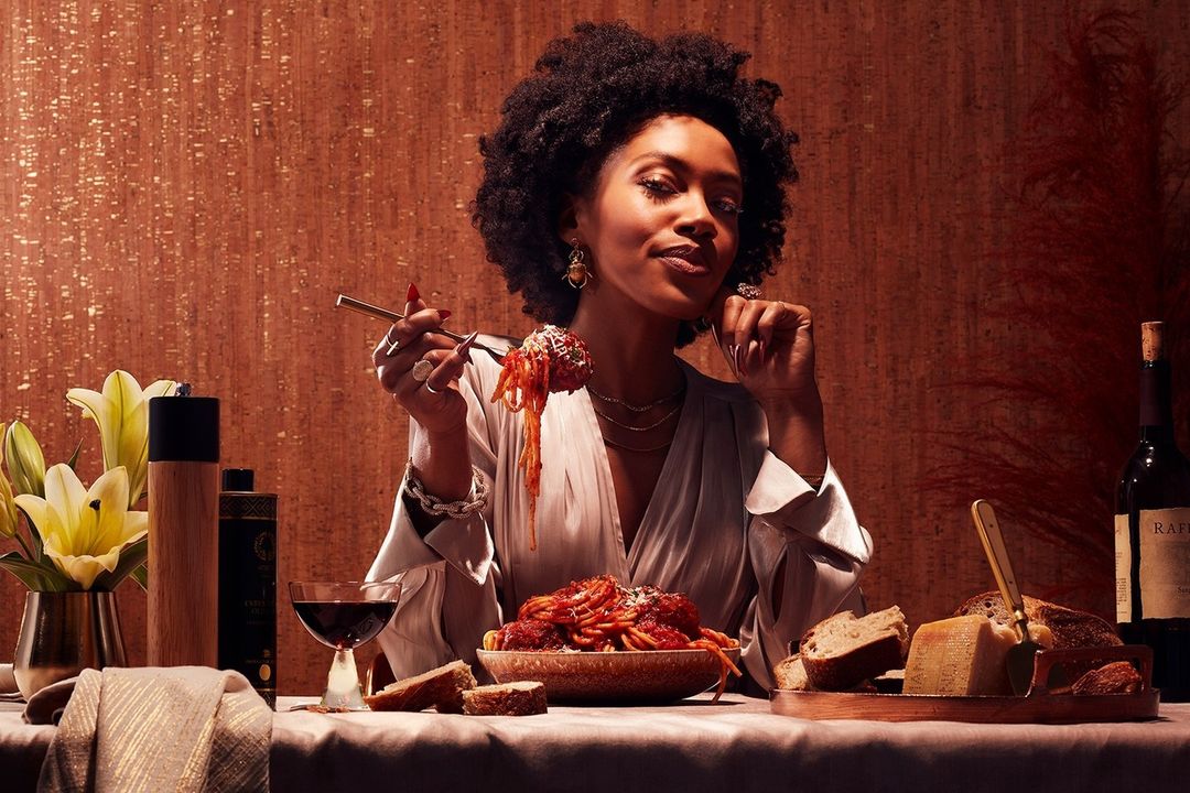 Elegant woman with afro hairstyle enjoys a plate of spaghetti with tomato sauce in a warmly lit indoor setting, wine, flowers.