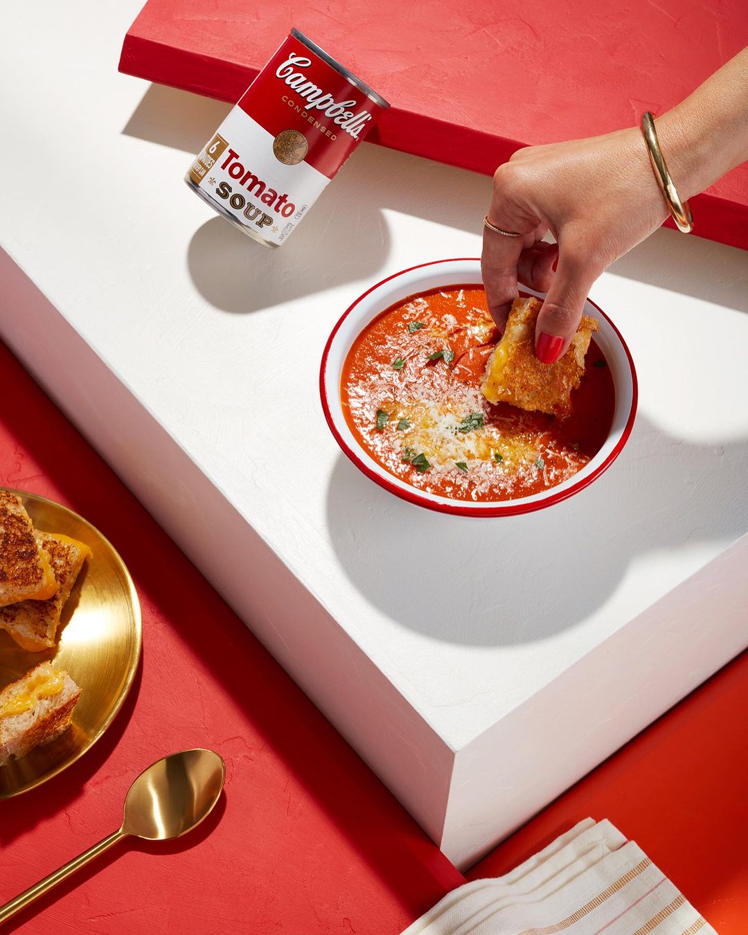 Tomato soup with grilled cheese crouton, Campbell's Tomato Soup can on a geometric white and red surface, golden spoon and gold bracelet.