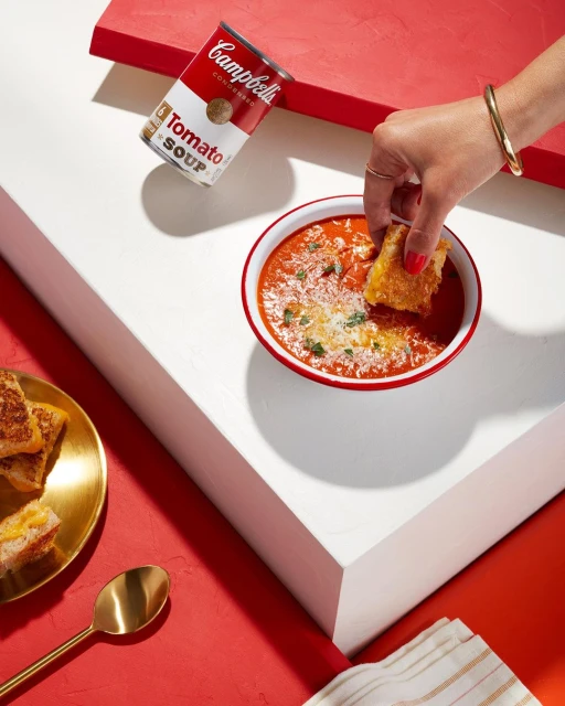 Tomato soup with grilled cheese crouton, Campbell's Tomato Soup can on a geometric white and red surface, golden spoon and gold bracelet.