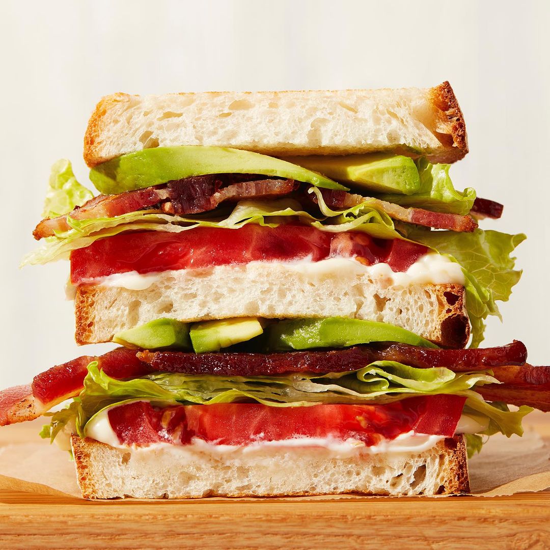 Stacked sandwich featuring bacon, lettuce, tomato, and avocado on toasted white bread, served on wooden board against a cream background.
