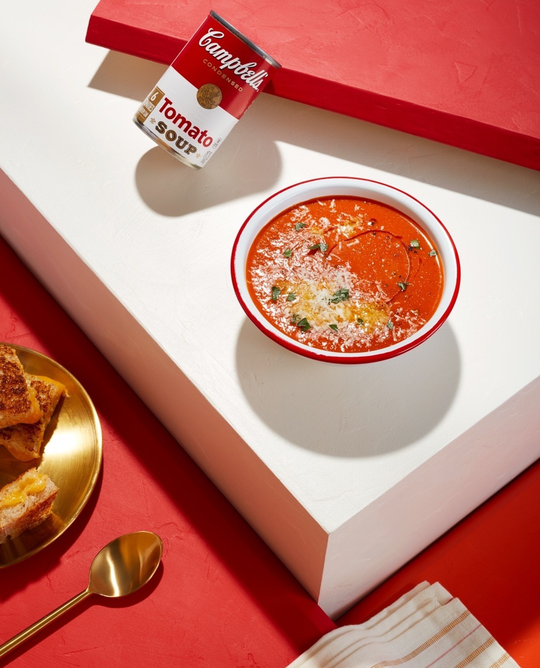 Still life shot of a bowl of tomato soup with a Campbell's soup can, grilled cheese sandwich, and gold spoon on white and red blocks.