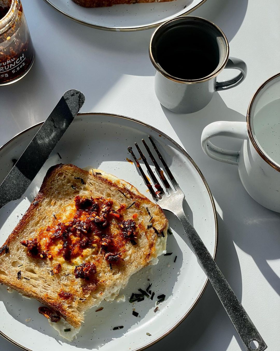 Overhead shot of toast topped with spicy sauce, a fork, knife, jar, and mugs on a white surface with natural sunlight.