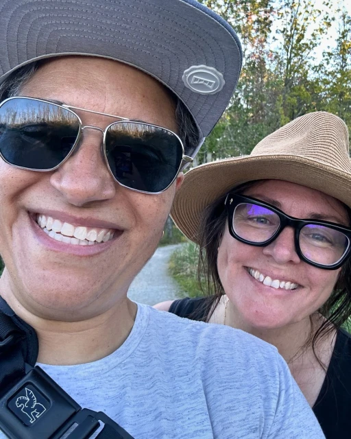Close-up of two smiling adult women outdoors, one wearing sunglasses and a baseball cap, the other in a hat and eyeglasses.