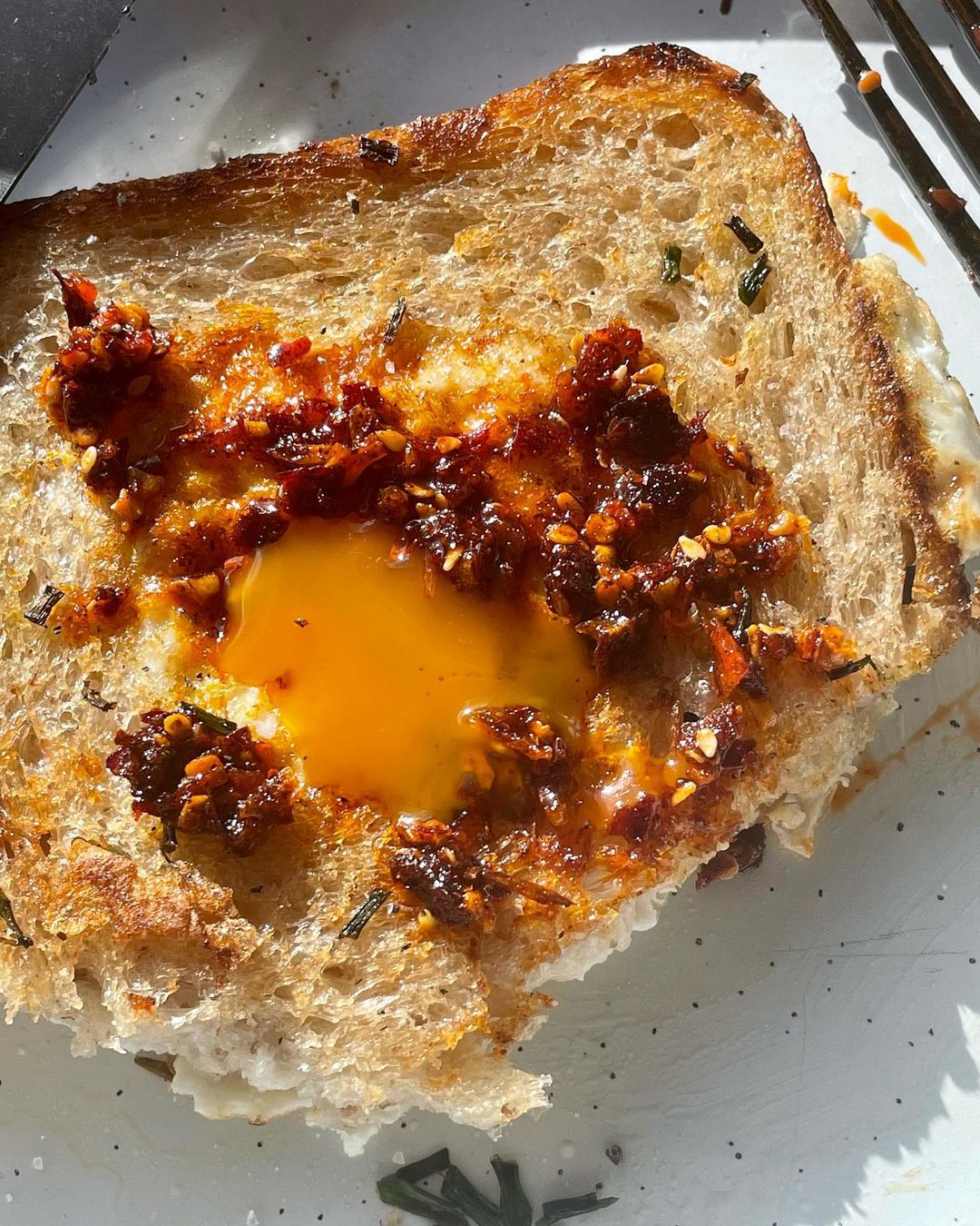 Toast with a fried egg in the center, topped with a reddish sauce and spices, sitting on a speckled plate, captured in bright, direct sunlight.