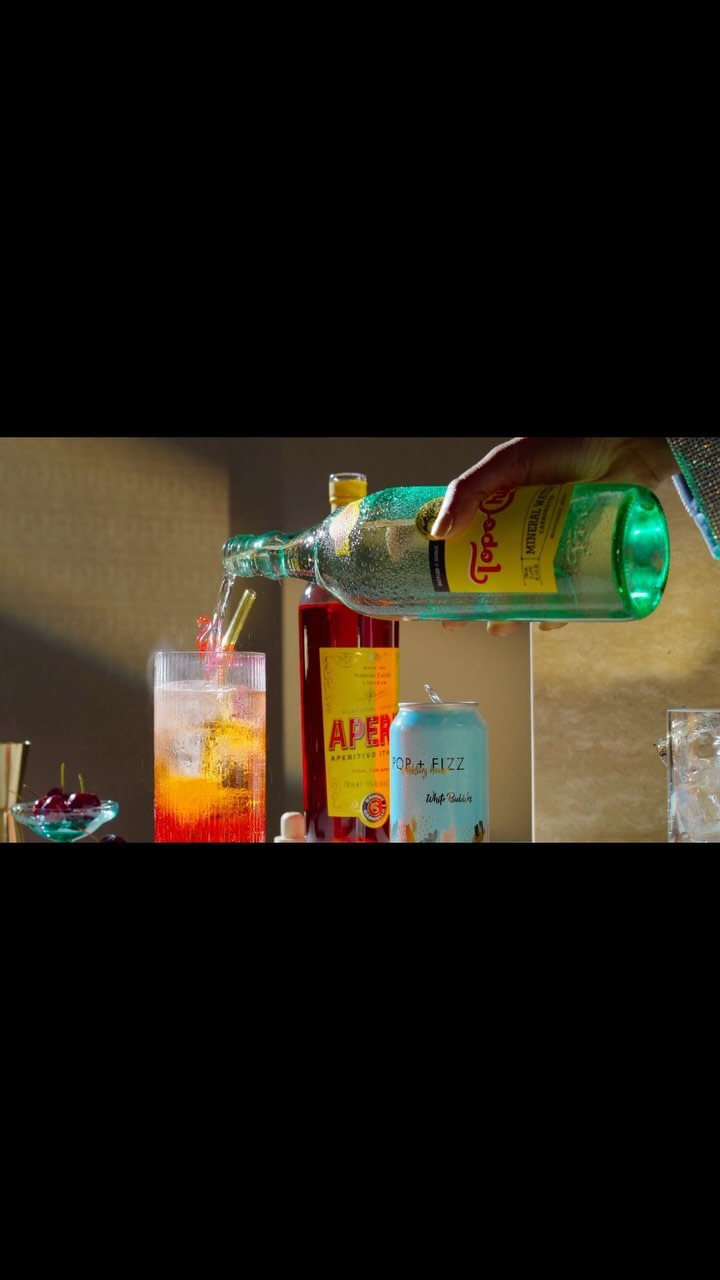 Person pouring Topo Chico mineral water into a glass with aperol and a straw next to a can and bottle of aperol on a counter.
