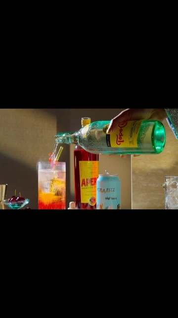 Person pouring Topo Chico mineral water into a glass with aperol and a straw next to a can and bottle of aperol on a counter.