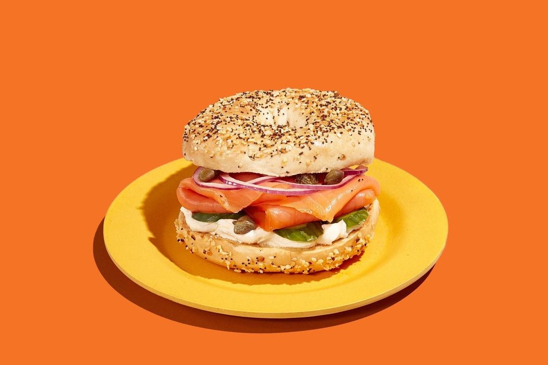 Everything bagel sandwich with smoked salmon, cream cheese, capers, and red onion served on a yellow plate against an orange backdrop.