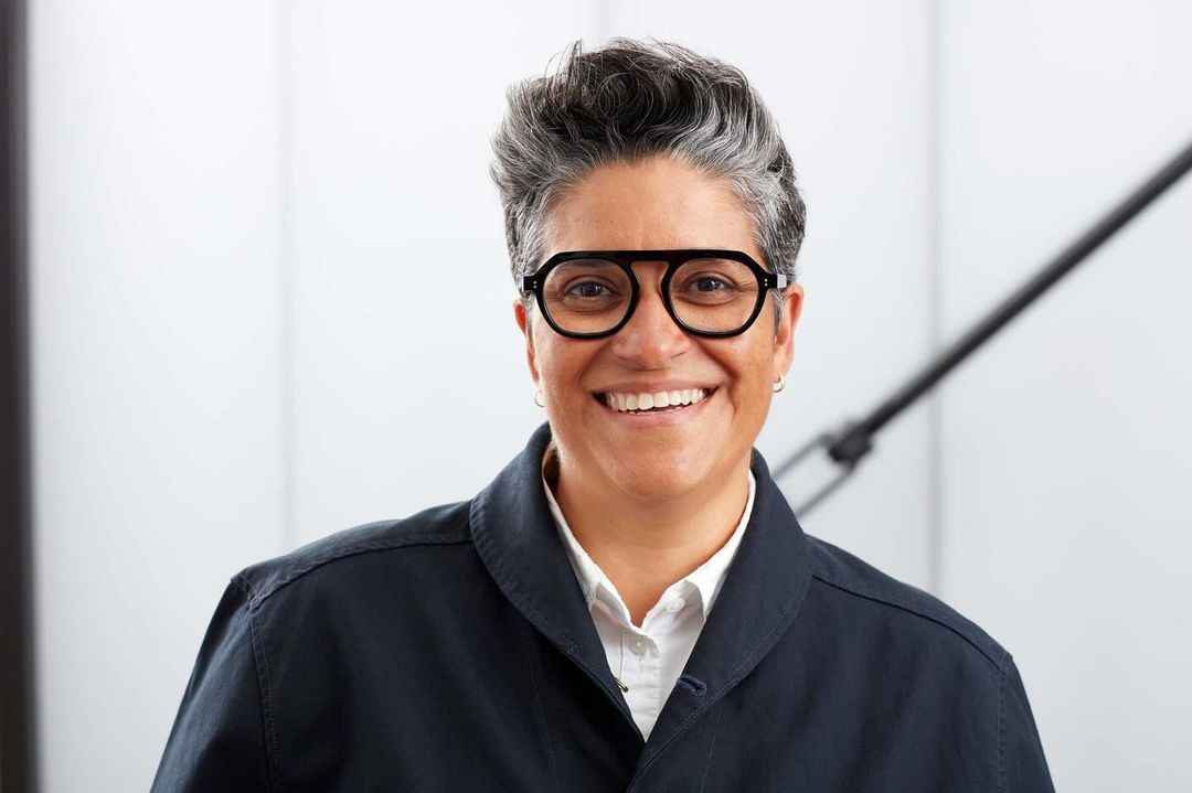 Portrait of a smiling person with short grey hair wearing black statement glasses and a dark button-up jacket over a white shirt.