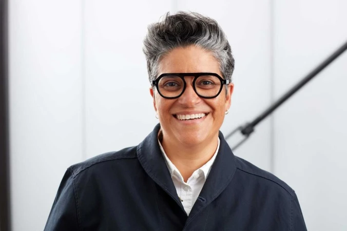 Portrait of a smiling person with short grey hair wearing black statement glasses and a dark button-up jacket over a white shirt.