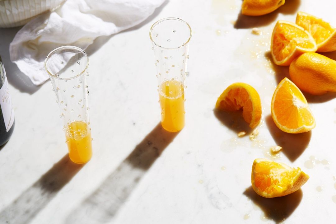 Two glasses of mimosa with fresh cut oranges. Orange slices and juice are scattered on a bright white marble surface.