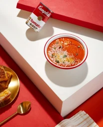 Bowl of tomato soup with grated cheese and herbs, accompanied by a Campbell's can and grilled cheese on a brass plate.