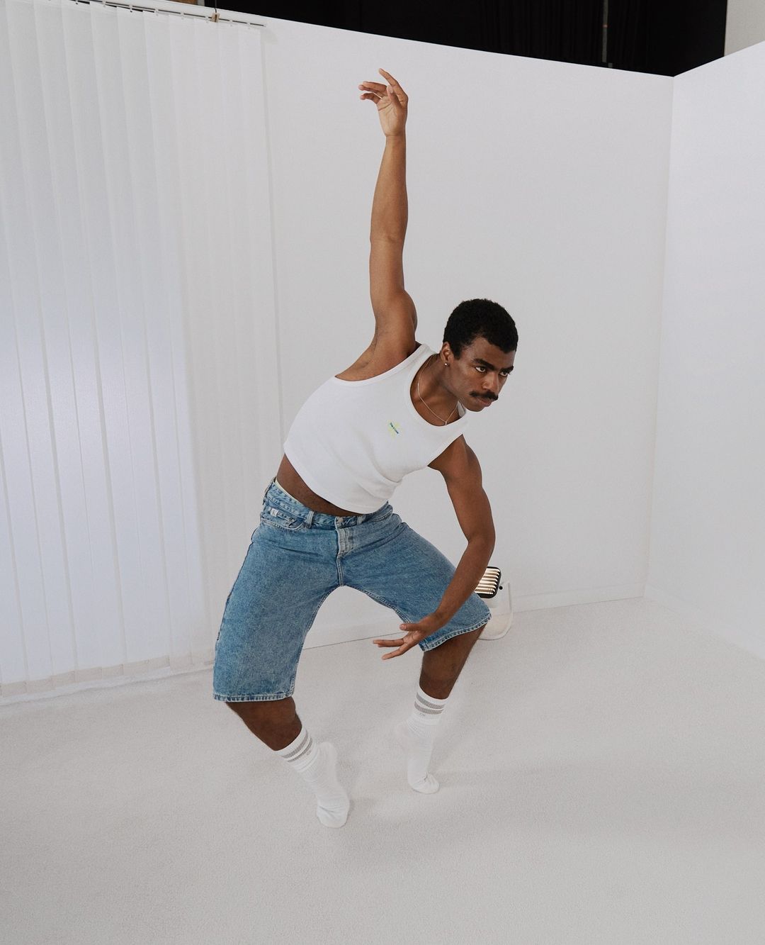 A man in a white tank top and denim shorts is captured in a dynamic, artistic pose in a bright studio with white walls.