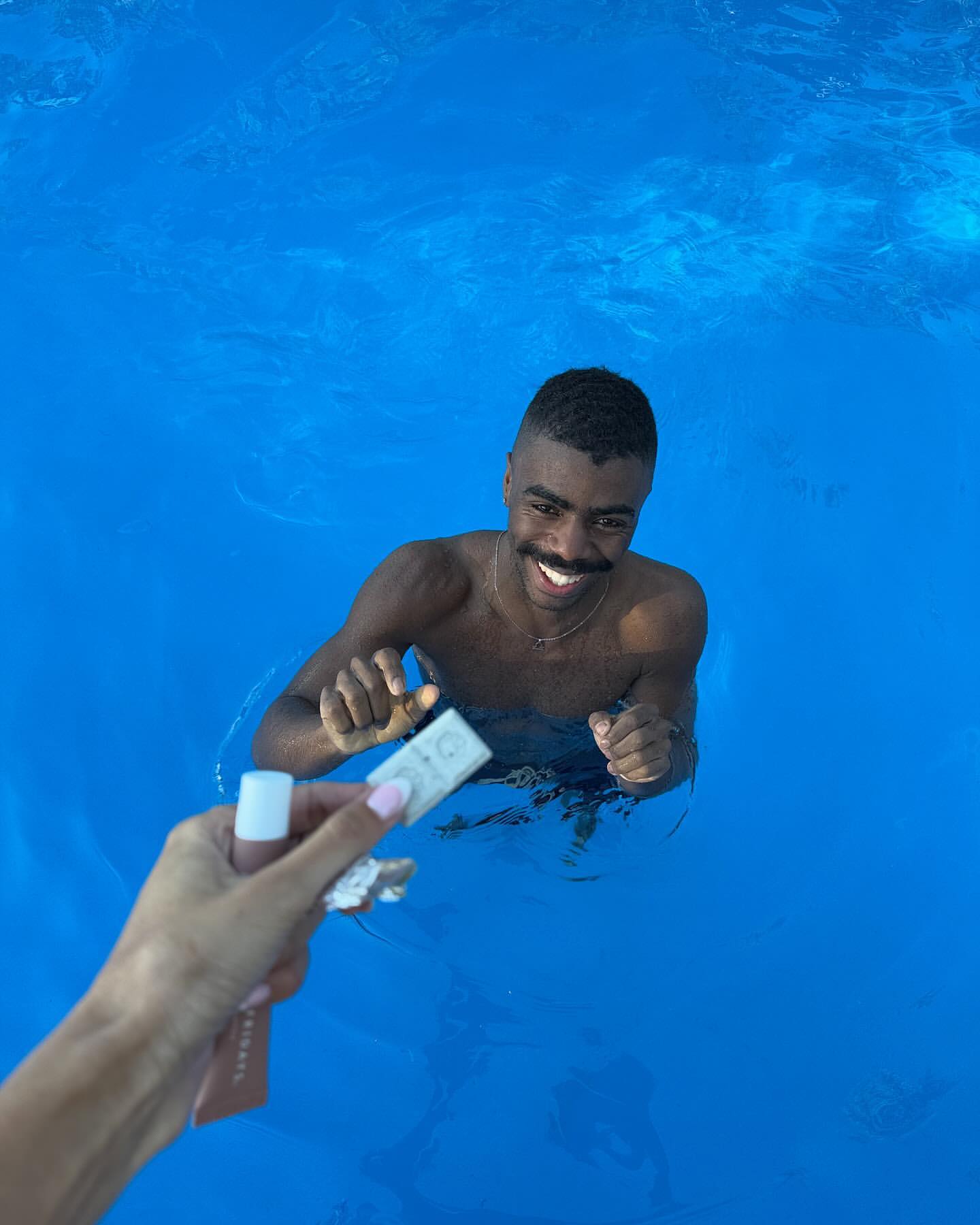 Smiling man in bright blue water receiving beauty products from outstretched arm, captured in medium shot under natural light.