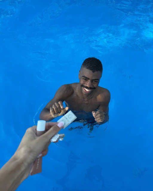 Smiling man in bright blue water receiving beauty products from outstretched arm, captured in medium shot under natural light.