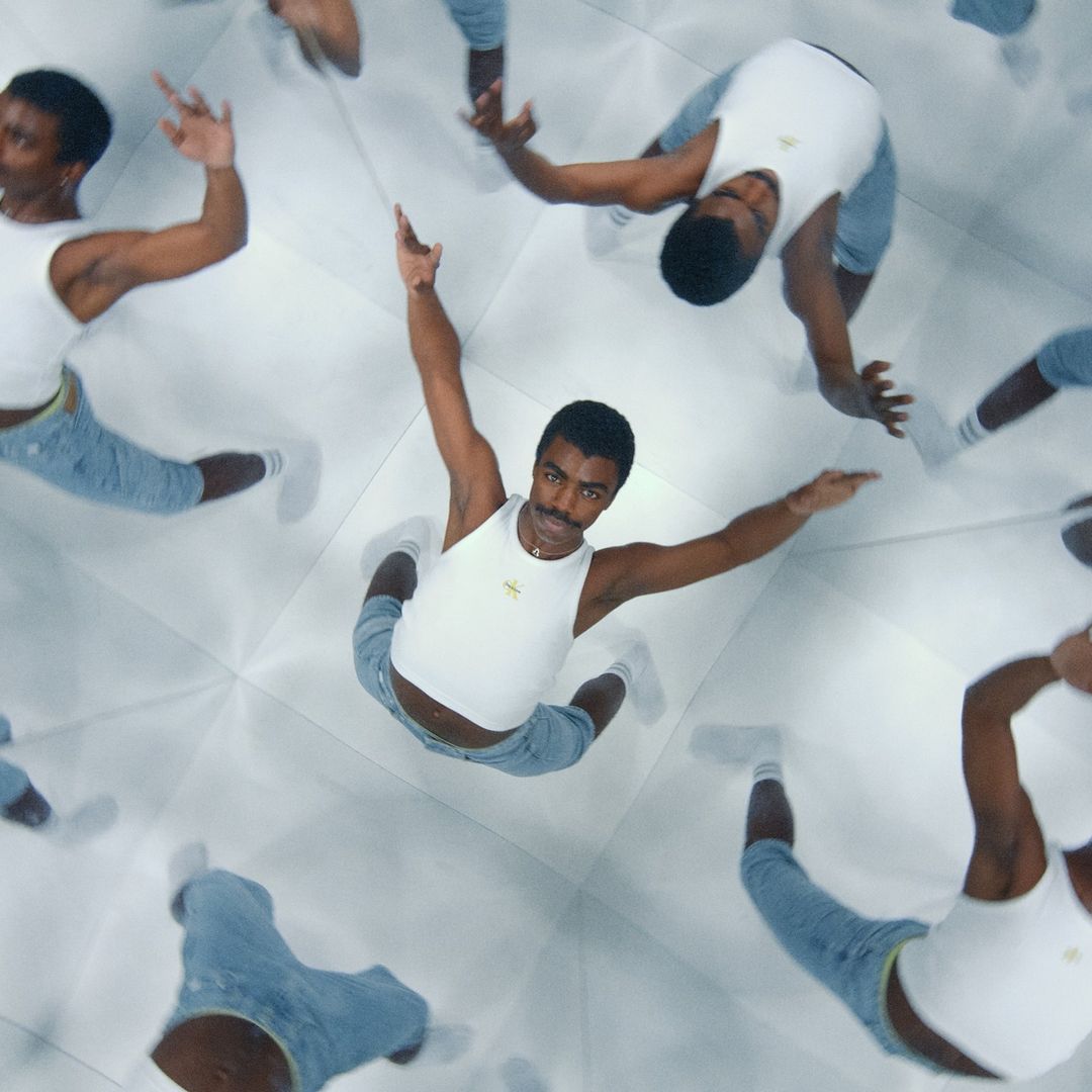 Man dances in kaleidoscopic mirror room, wearing white tank top and light blue jeans, creating multiple reflections and dynamic poses.