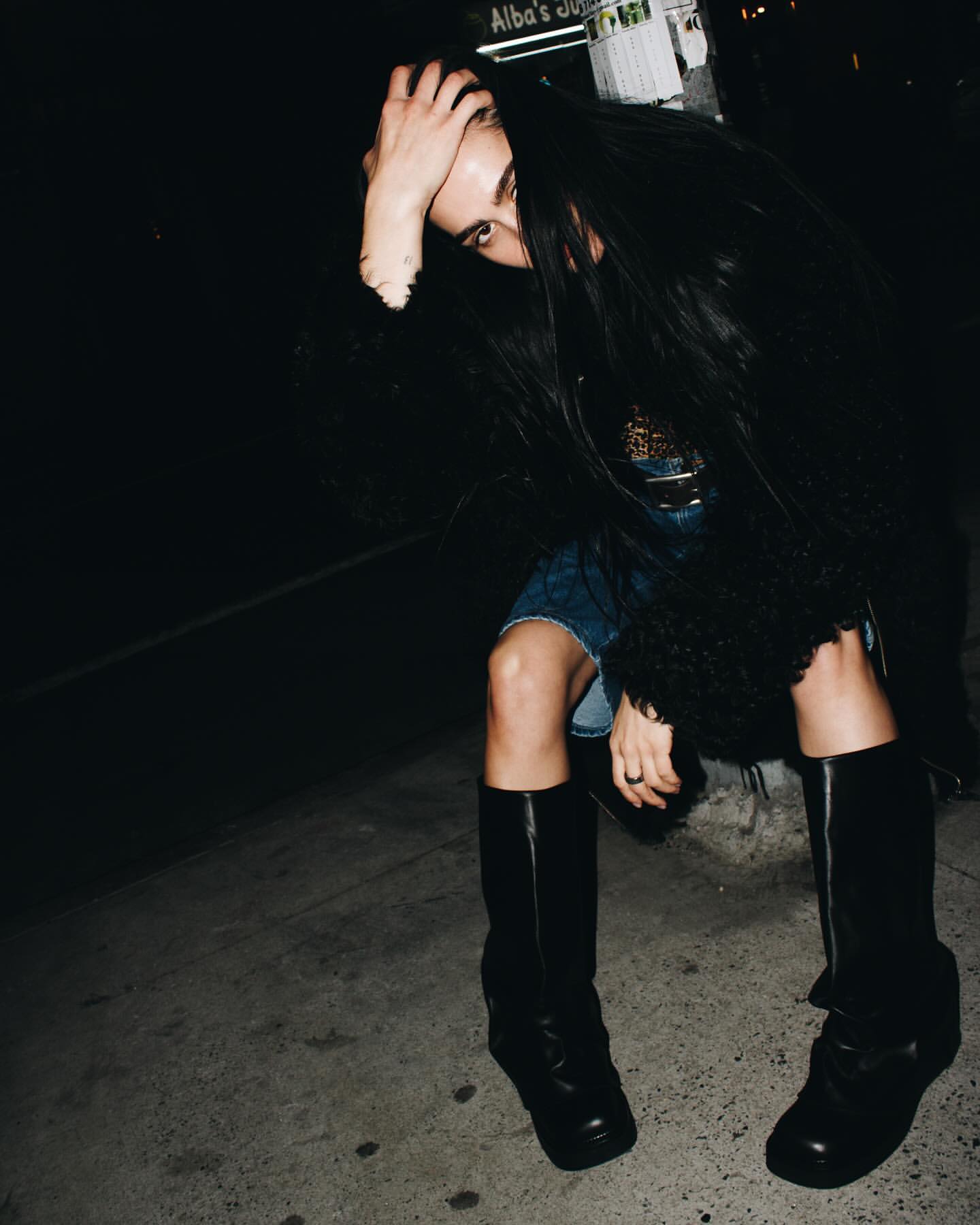 Woman with long dark hair covering her face sitting on a New York street wearing tall black boots.
