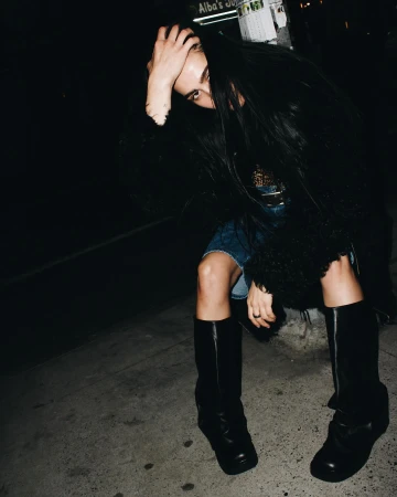 Woman with long dark hair covering her face sitting on a New York street wearing tall black boots.