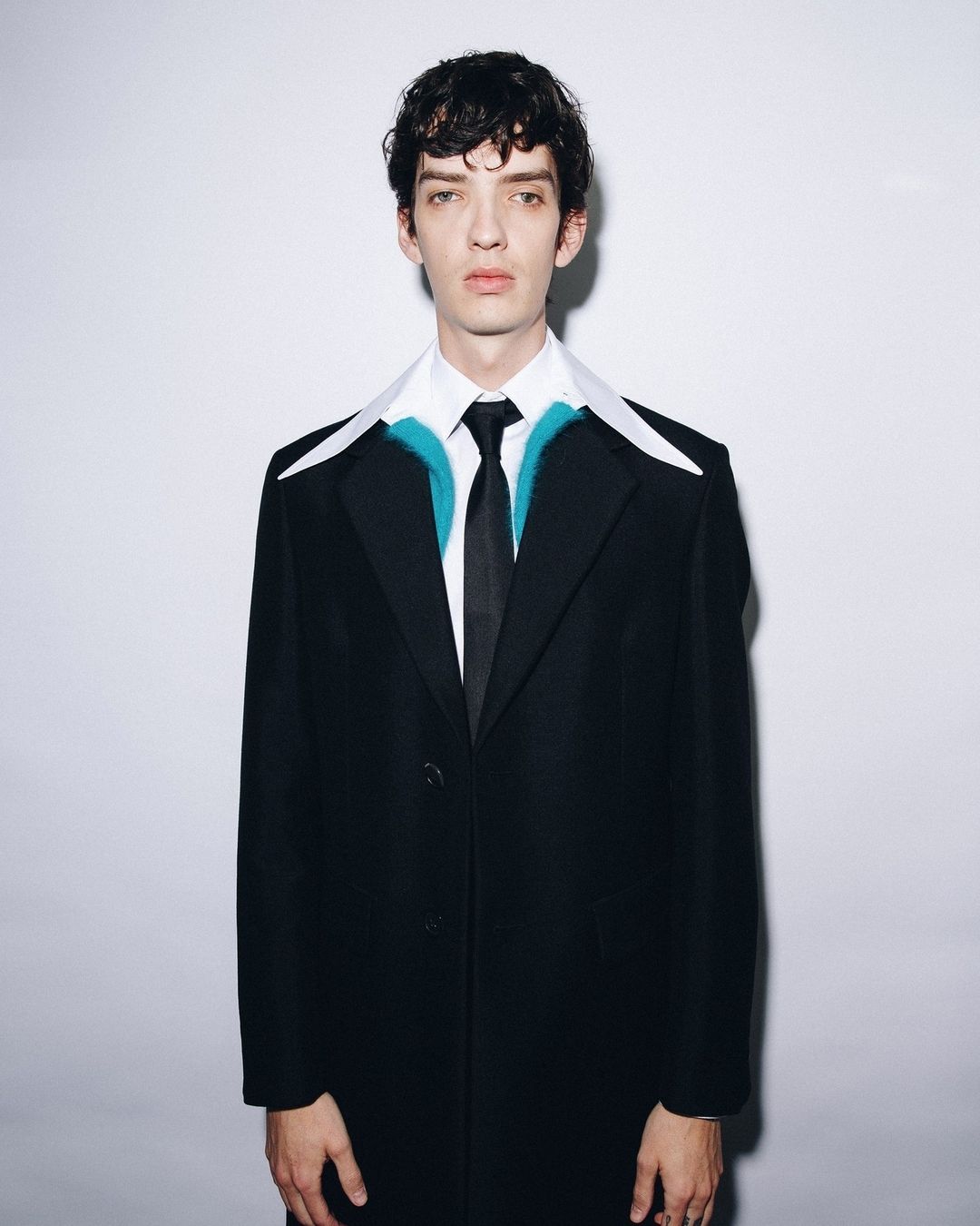 Man in a black coat, white shirt, and black tie stands against a white background in a studio portrait; fashion and style.