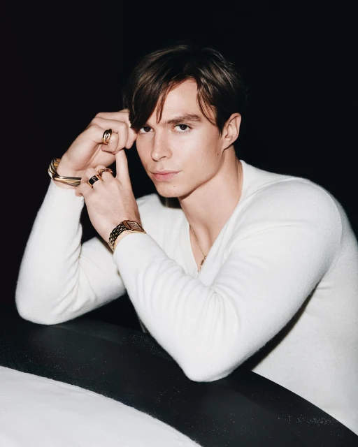 Portrait of a young adult man in a white sweater, wearing gold rings and bracelet, posing against a black and white background.