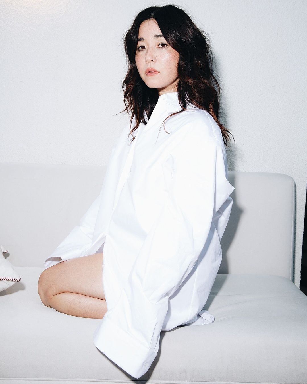 Portrait of a woman with dark wavy hair, wearing an oversized white shirt, sitting on a white sofa in a bright studio.