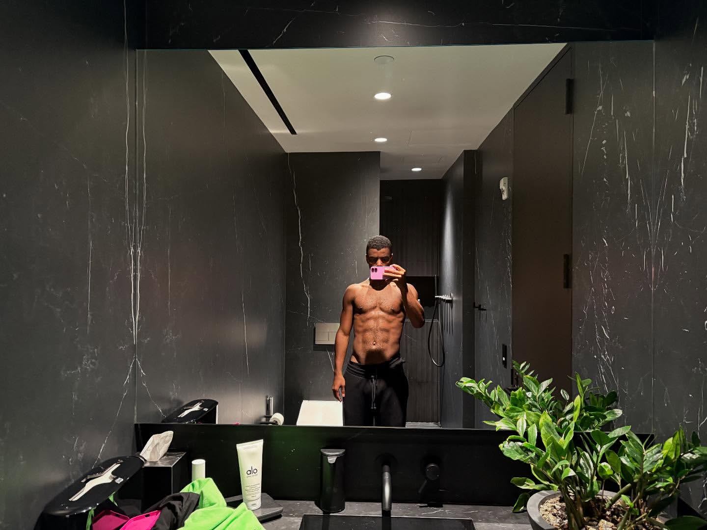 Athletic man takes a mirror selfie in a modern bathroom with dark marble walls, holding a pink phone, showcasing his physique and healthy lifestyle.