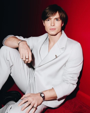 Young man in a light grey suit seated against a vibrant red backdrop, photographed by Tyrell Hampton.