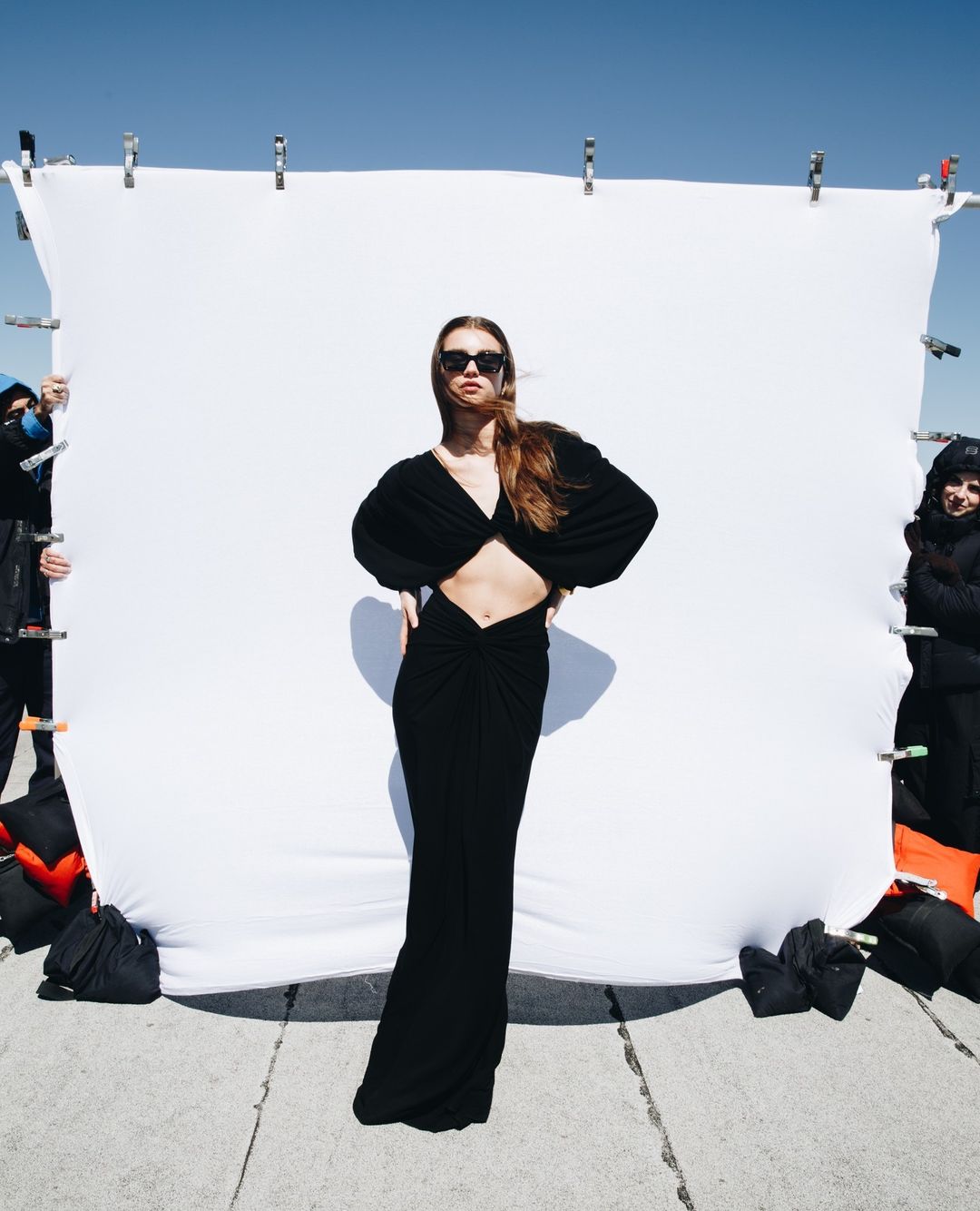 A fair-skinned woman with sunglasses poses in a black dress against a white backdrop outdoors during a fashion photoshoot.