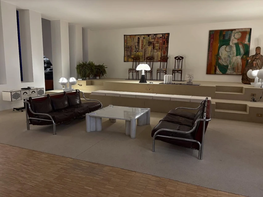 Living room with vintage brown leather couches, white marble coffee table, decorative art, and tiered platform in neutral, indoor setting.